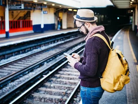 Pejalan dengan ransel kuning dan topi menunggu kereta sambil melihat ponsel di peron stasiun Richmond, Inggris