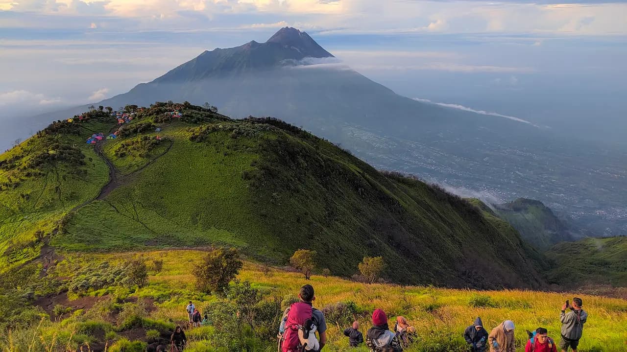 Pendaki berjalan di sabana jalur Selo Gunung Merbabu dengan tenda di punggungan dan Gunung Merapi di latar belakang