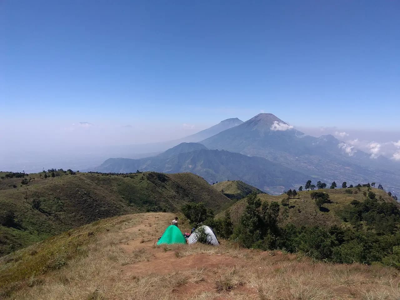 Punggungan berumput di puncak Gunung Prau dengan dua tenda, Gunung Sindoro dan Sumbing tampak di kejauhan