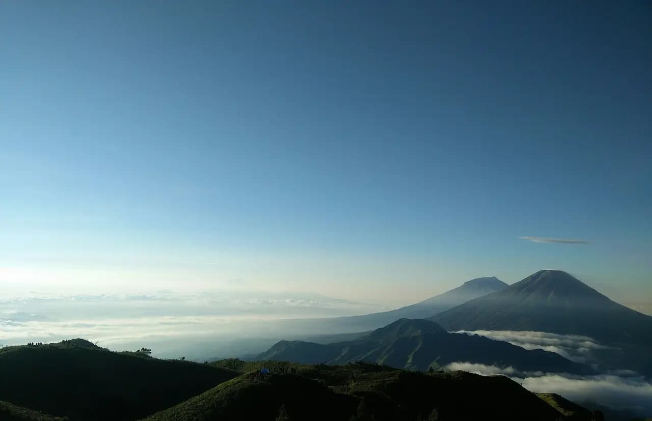 Bukit-bukit berumput Gunung Prau pada pagi hari dengan Gunung Sindoro dan Sumbing di atas lautan awan