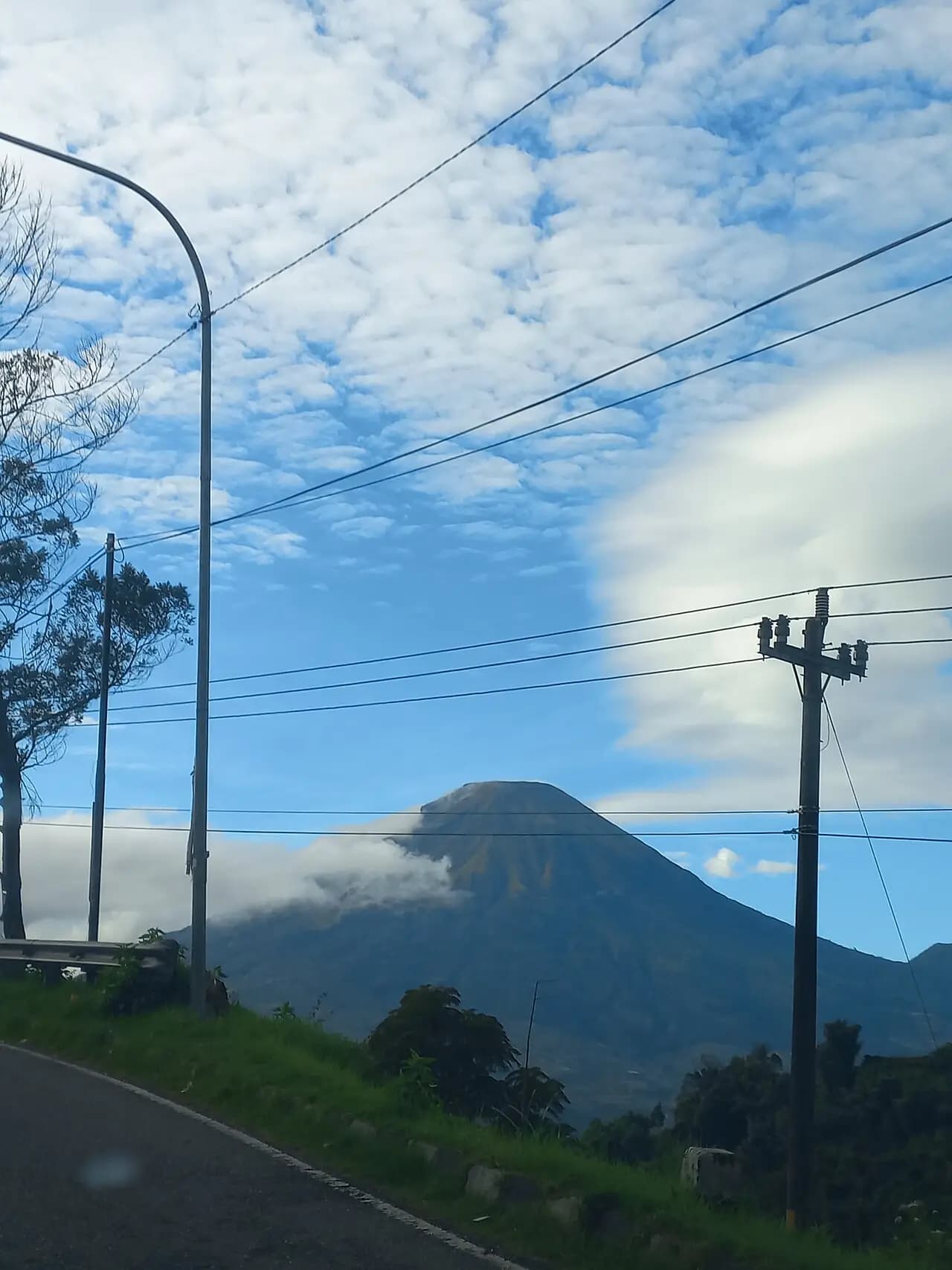 Kerucut Gunung Sindoro sebagian tertutup awan, dilihat dari tepi jalan Wonosobo-Dieng dengan tiang listrik di depan