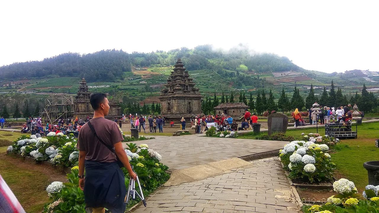 Kompleks Candi Arjuna dengan banyak pengunjung berkelompok, taman bunga di depan dan bukit bertingkat di belakang