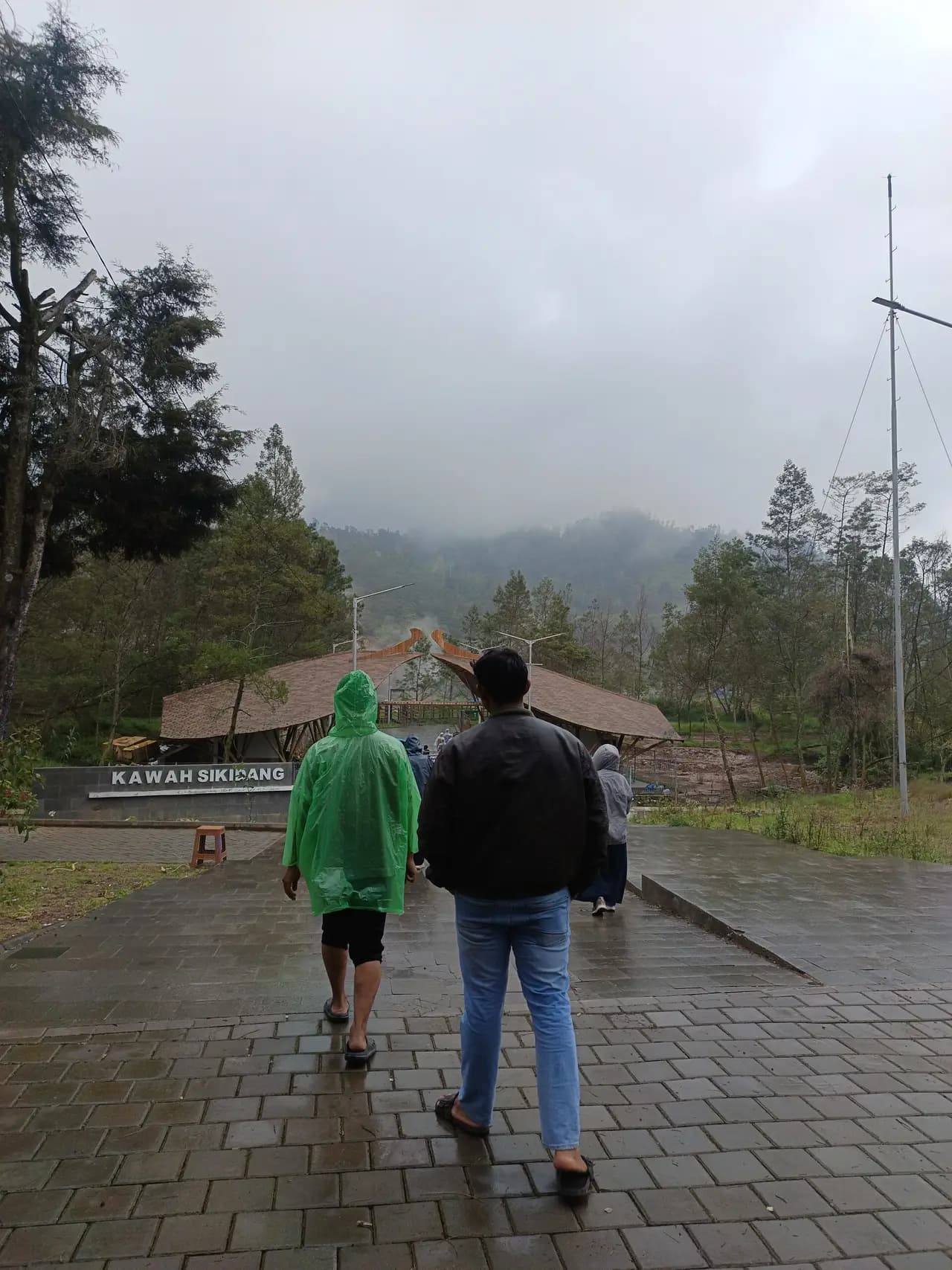 Pengunjung, satu di antaranya berjas hujan hijau, berjalan di jalan batu basah menuju bangunan beratap kayu di pintu masuk Kawah Sikidang; papan nama Kawah Sikidang di dinding batu kiri, uap kawah di belakang