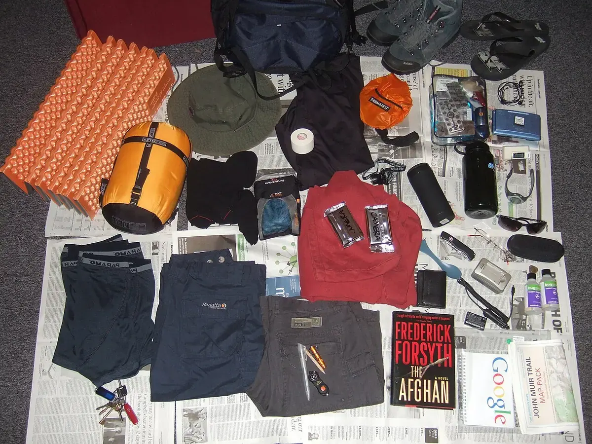 Peralatan pendakian ditata di lantai: matras busa oranye, sleeping bag kuning, topi, headlamp, botol minum, kotak obat, dan pakaian