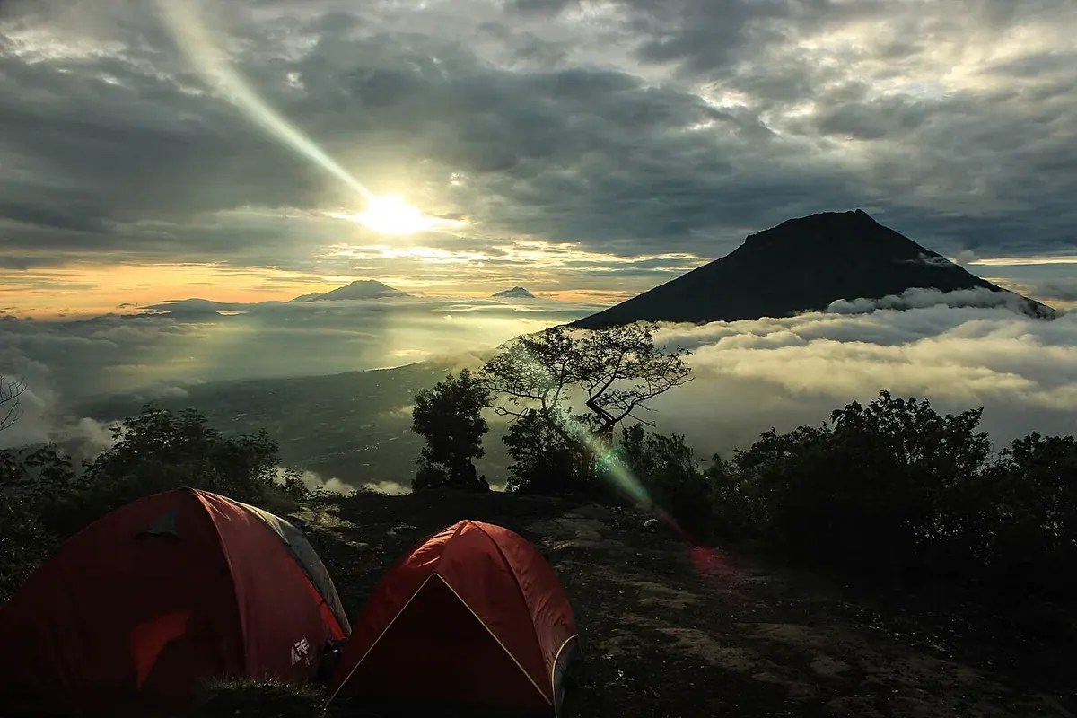 Dua tenda dome merah berdiri di lereng Gunung Sindoro pada pagi berawan, dengan lautan awan dan kerucut gunung di latar belakang