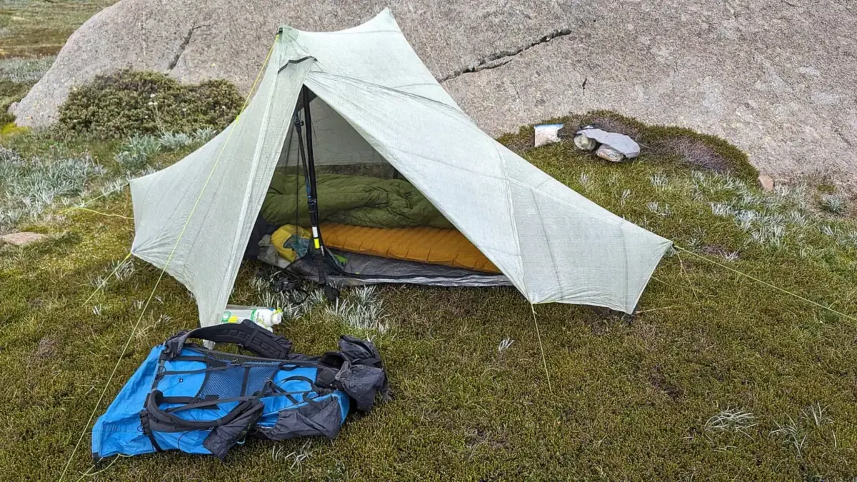 Tenda ultralight dengan sleeping bag hijau dan matras oranye di dalamnya, serta ransel biru di depan tenda