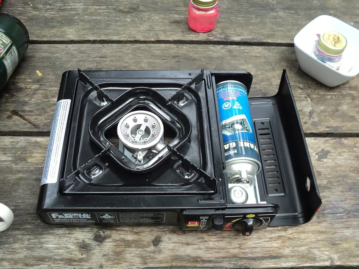 Kompor gas portable hitam dengan penutup samping terbuka memperlihatkan tabung gas butana yang terpasang