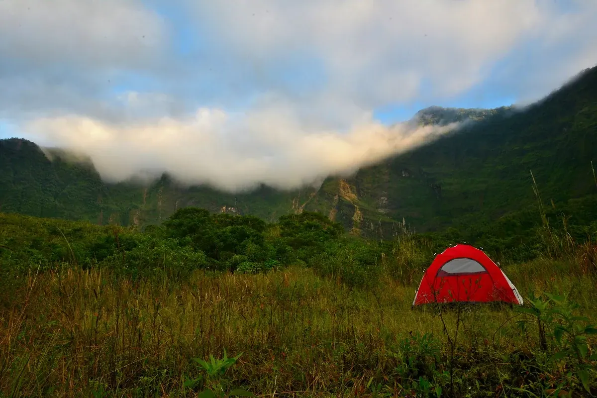 Tenda dome merah berdiri di padang rumput dalam kaldera Gunung Galunggung, dengan tebing hijau berselimut awan di belakangnya