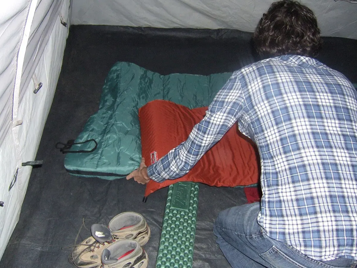 Seseorang menata sleeping bag hijau dan matras oranye di dalam tenda, dengan sepasang sepatu di sampingnya