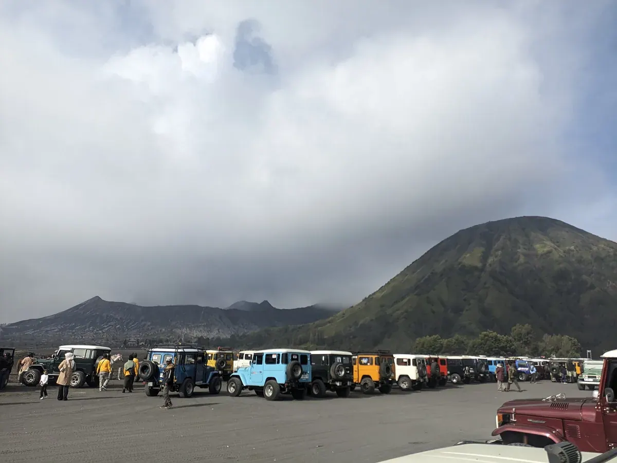 Deretan jip wisata berwarna-warni terparkir di hamparan pasir dengan gunung hijau di kanan dan awan tebal di atasnya
