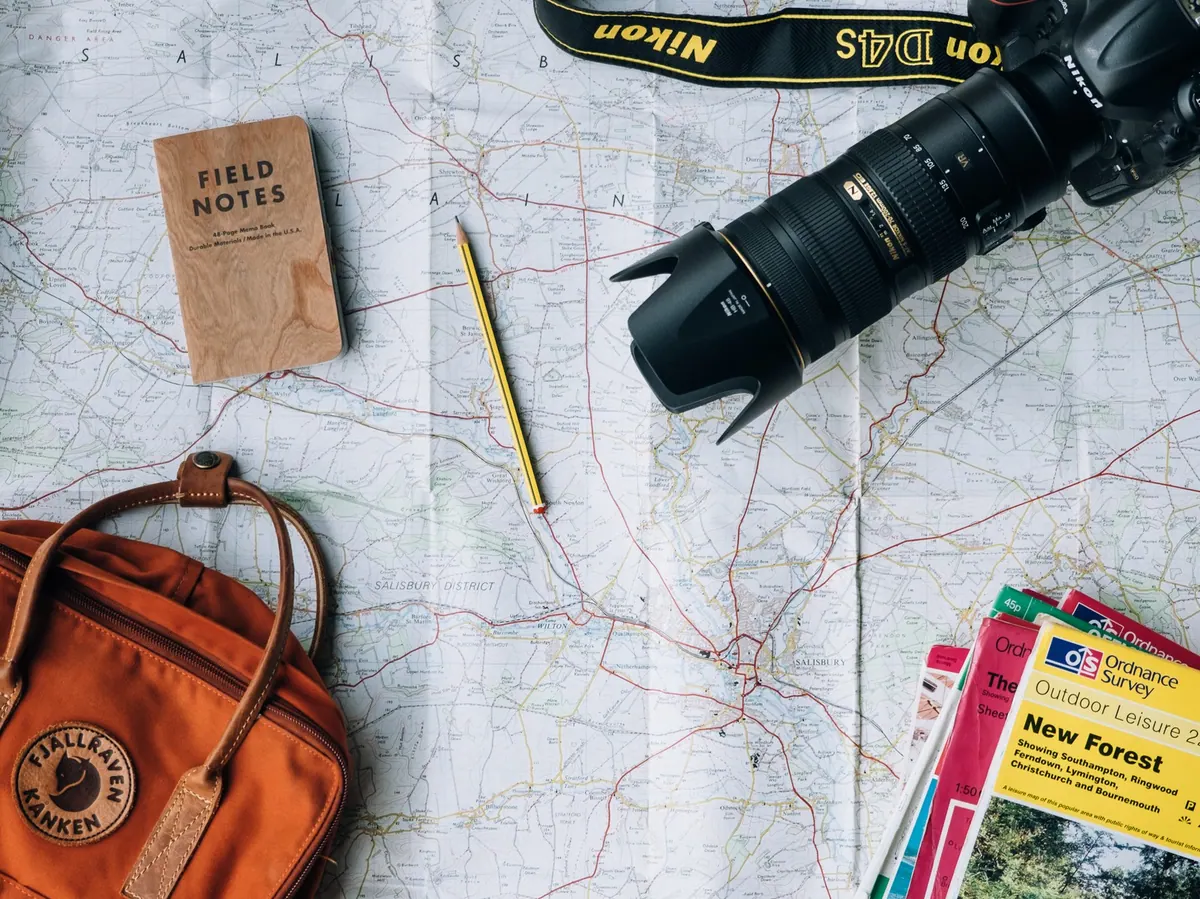 Peta jalan terbentang di meja bersama buku catatan, pensil, kamera, tas, dan brosur wisata untuk menyusun rencana perjalanan