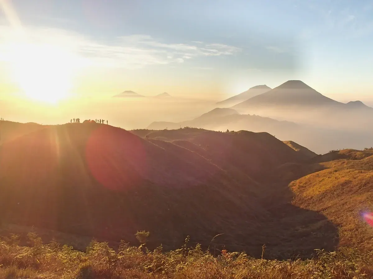 Matahari terbit dari punggungan Gunung Prau, dengan siluet pendaki di puncak bukit dan deretan gunung api Jawa Tengah di kejauhan