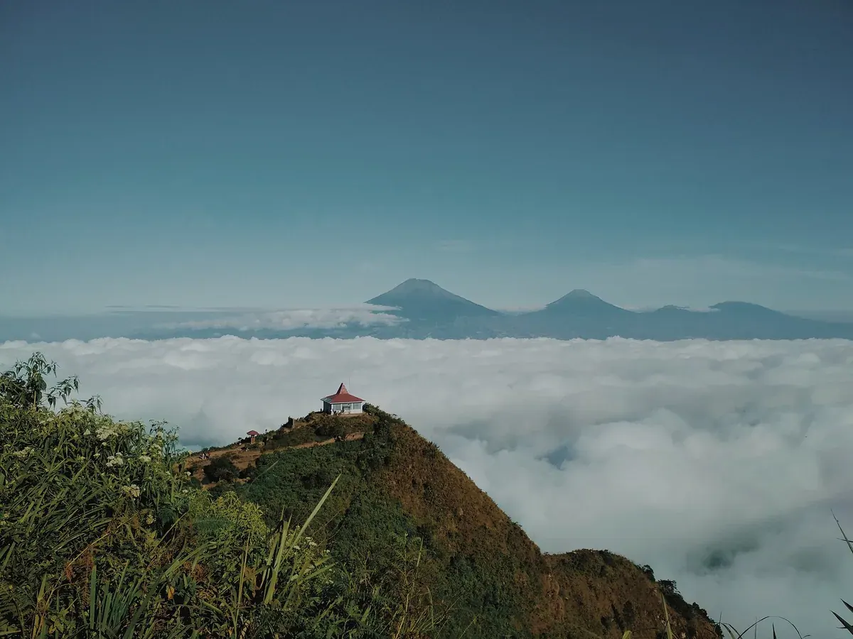 Punggungan puncak Gunung Andong dengan bangunan beratap merah di atas lautan awan