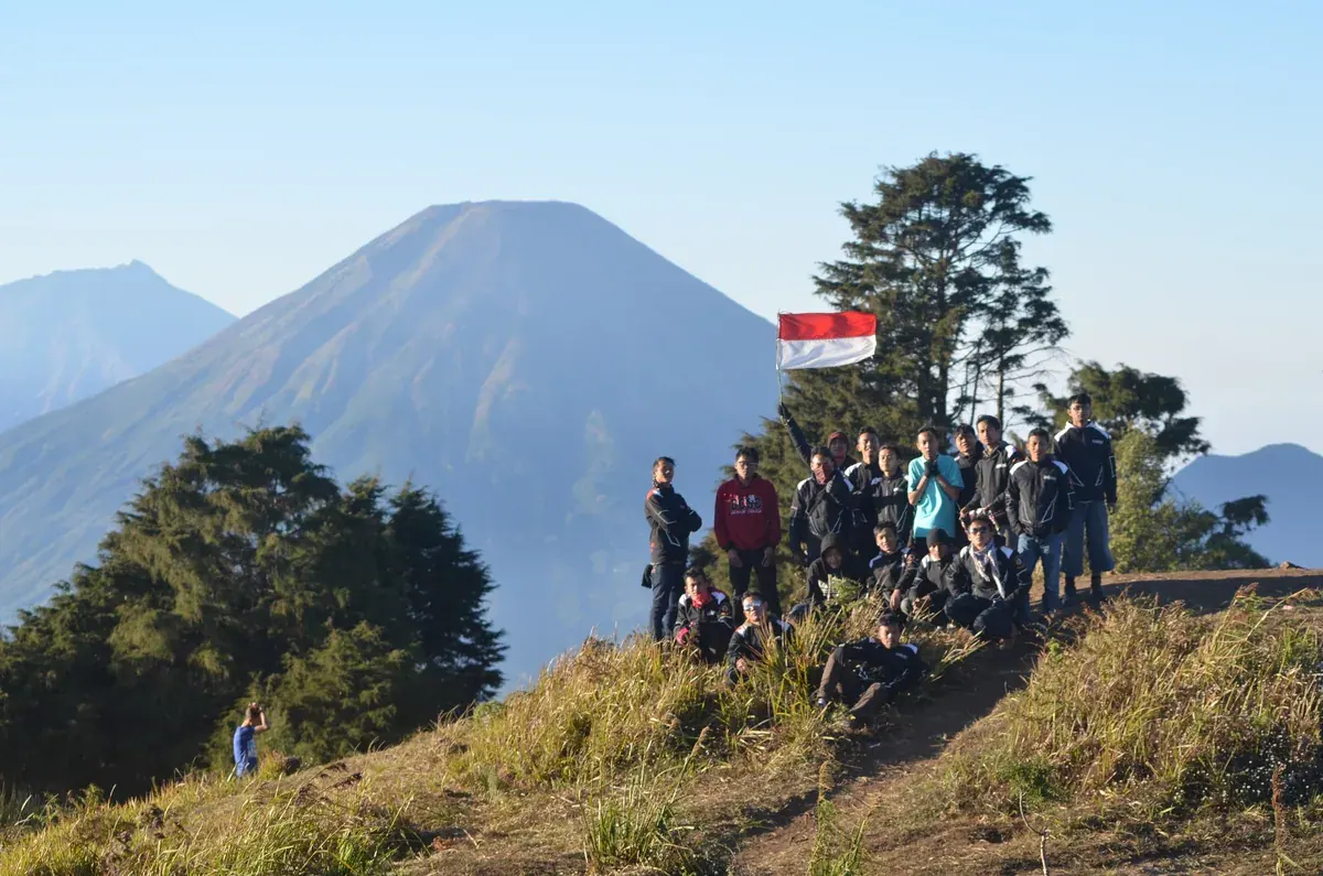 Rombongan pendaki berkumpul di punggungan berumput Gunung Prau, Dieng, dengan gunung berbentuk kerucut di latar belakang