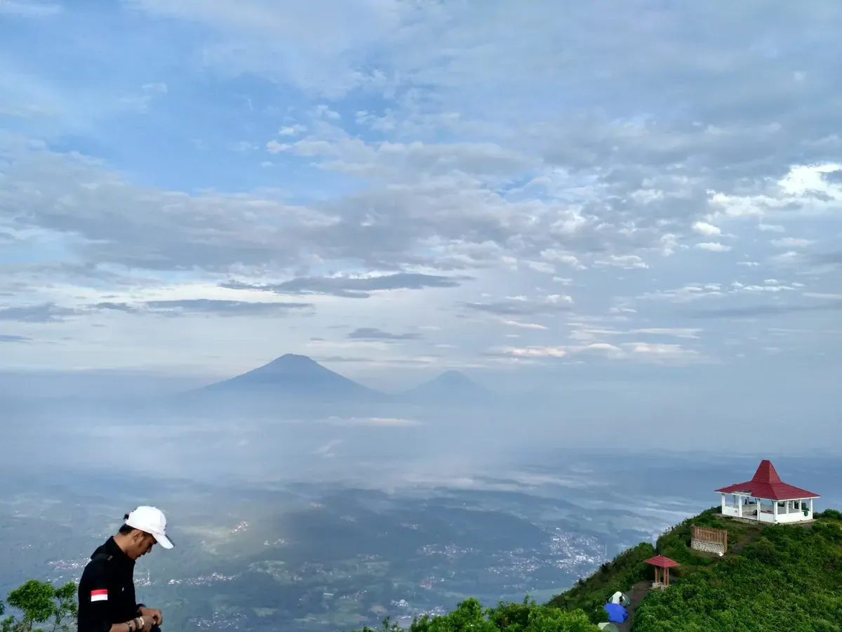 Pendaki berdiri di puncak Gunung Andong dengan gazebo beratap merah, beberapa tenda, dan lautan awan di kejauhan