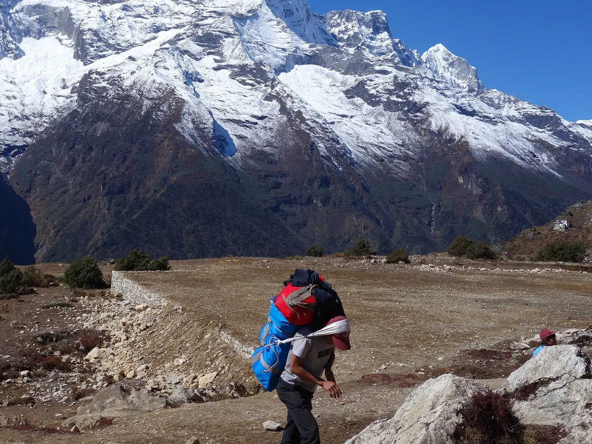 Porter memikul tas besar dengan tali pengikat di kepala di jalur pendakian kawasan Everest, Nepal, berlatar pegunungan bersalju