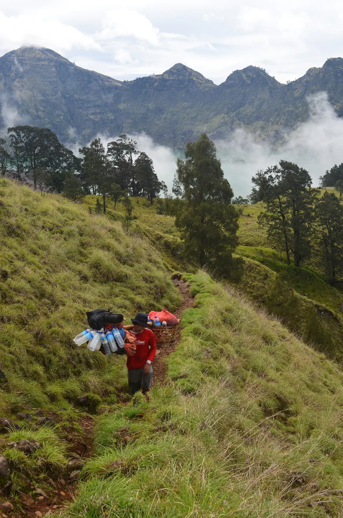 Porter berbaju merah memikul dua keranjang berisi botol air, tas, dan logistik rombongan di jalur berumput Gunung Rinjani