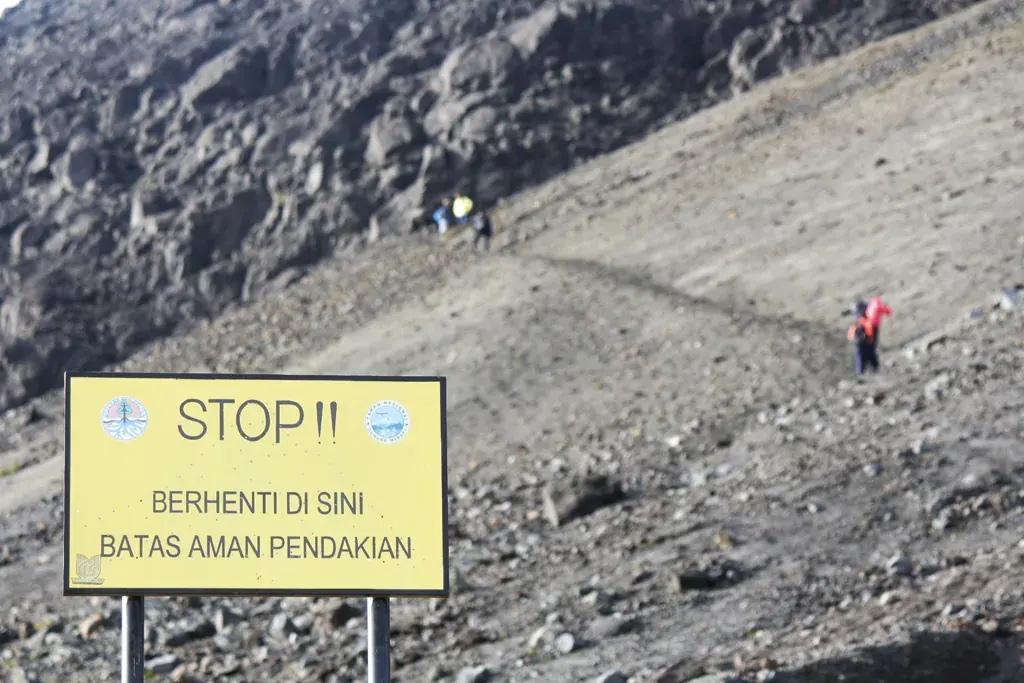 Papan kuning bertuliskan STOP, berhenti di sini, batas aman pendakian, di lereng berpasir Pasar Bubrah Gunung Merapi, dengan beberapa pendaki kecil di kejauhan