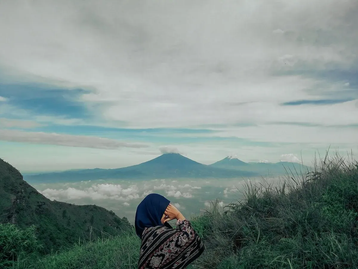 Pendaki berkerudung membelakangi kamera di lereng berumput jalur Gunung Andong via Sawit, memandang deretan gunung di atas lautan awan
