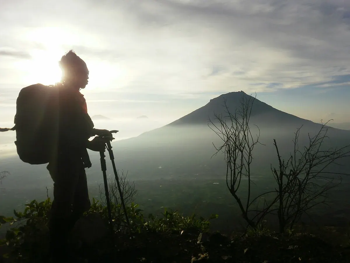 Siluet pendaki dengan ransel dan dua tongkat trekking di lereng Gunung Sindoro pada pagi hari, dengan Gunung Sumbing di latar belakang