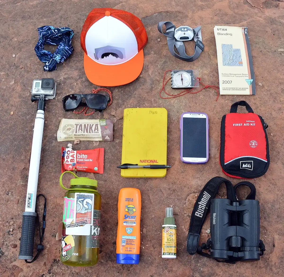 Barang bawaan hiking yang ditata di atas batu merah: bandana, topi, lampu kepala, kompas, peta, kacamata hitam, camilan, buku catatan, ponsel, kotak P3K, botol minum, tabir surya, semprotan antinyamuk, teropong, dan kamera dengan tongkatnya
