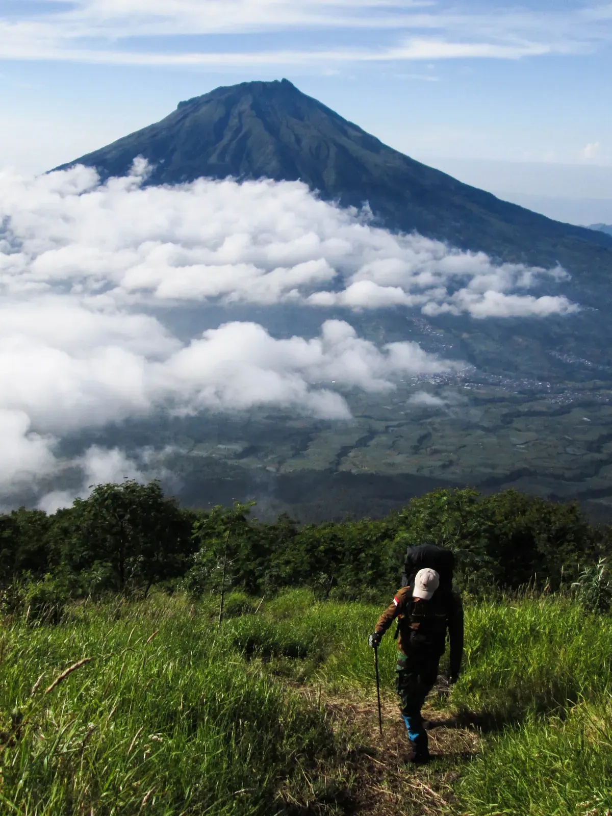 Pendaki bertopi dengan tongkat pendakian dan ransel menapaki jalur berumput di Gunung Sindoro, dengan kerucut Gunung Sumbing di belakang lautan awan