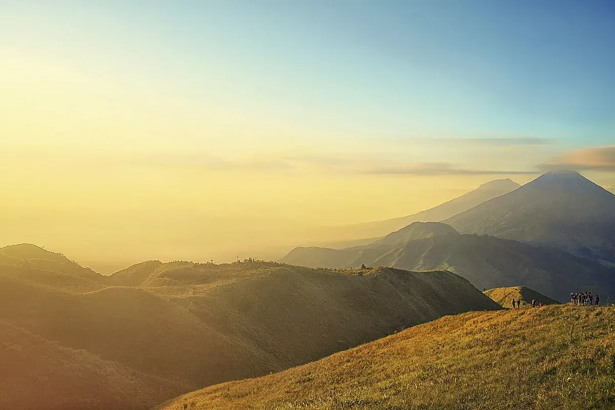 Punggungan berumput Gunung Prau di pagi hari dengan rombongan pendaki kecil di kejauhan dan deretan gunung berselimut kabut
